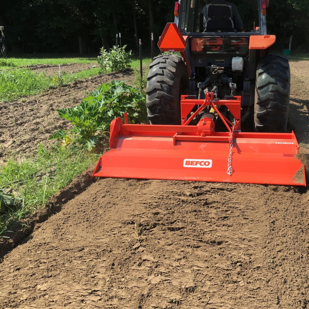 BEFCO T70 Rotary Tiller, red, angled view with metal tines and attached to a tractor.