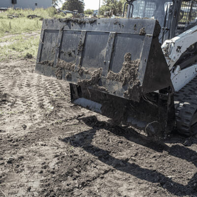 Skid Steer 4-in-1 Bucket by Prime Attachments, angled view, black with a dirt-filled scoop.