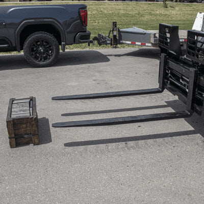 Prime Attachments hydraulic adjustable forks in black, angled view with extended tines beside a wooden crate.