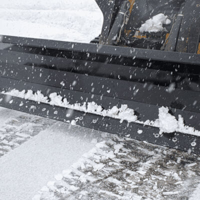 Skid Steer Snow Plow Blade by Prime Attachments, black, angled view with snow accumulation.