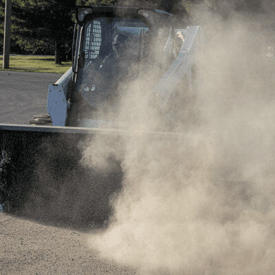 Prime Attachments Skid Steer Sweeper Attachment front view, gray with rotating broom and dust cloud.